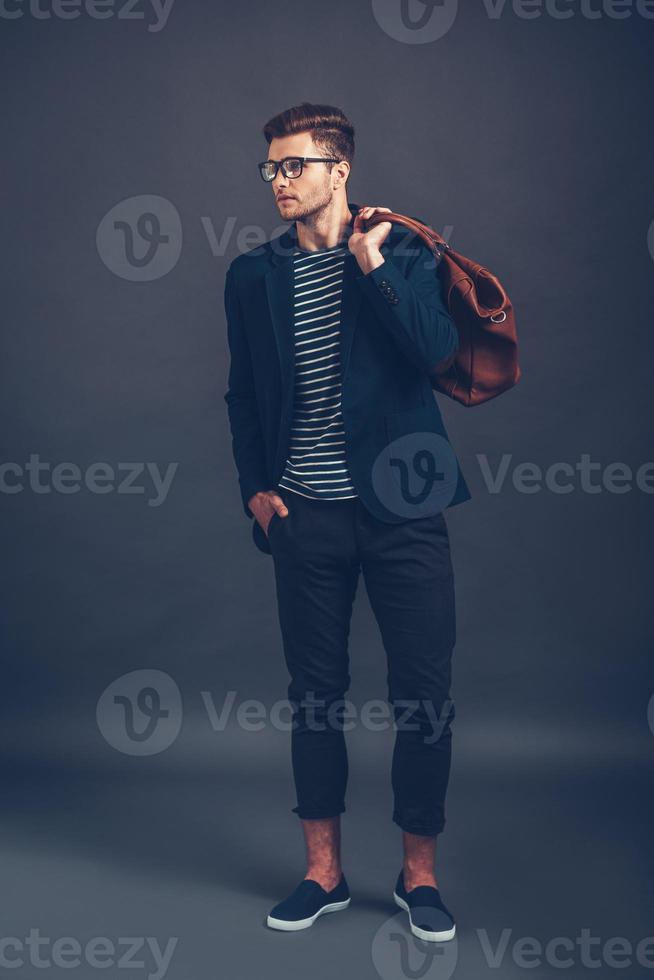 Confident in his style. Full length of confident young handsome man in glasses holding bag and looking away while standing against grey background photo