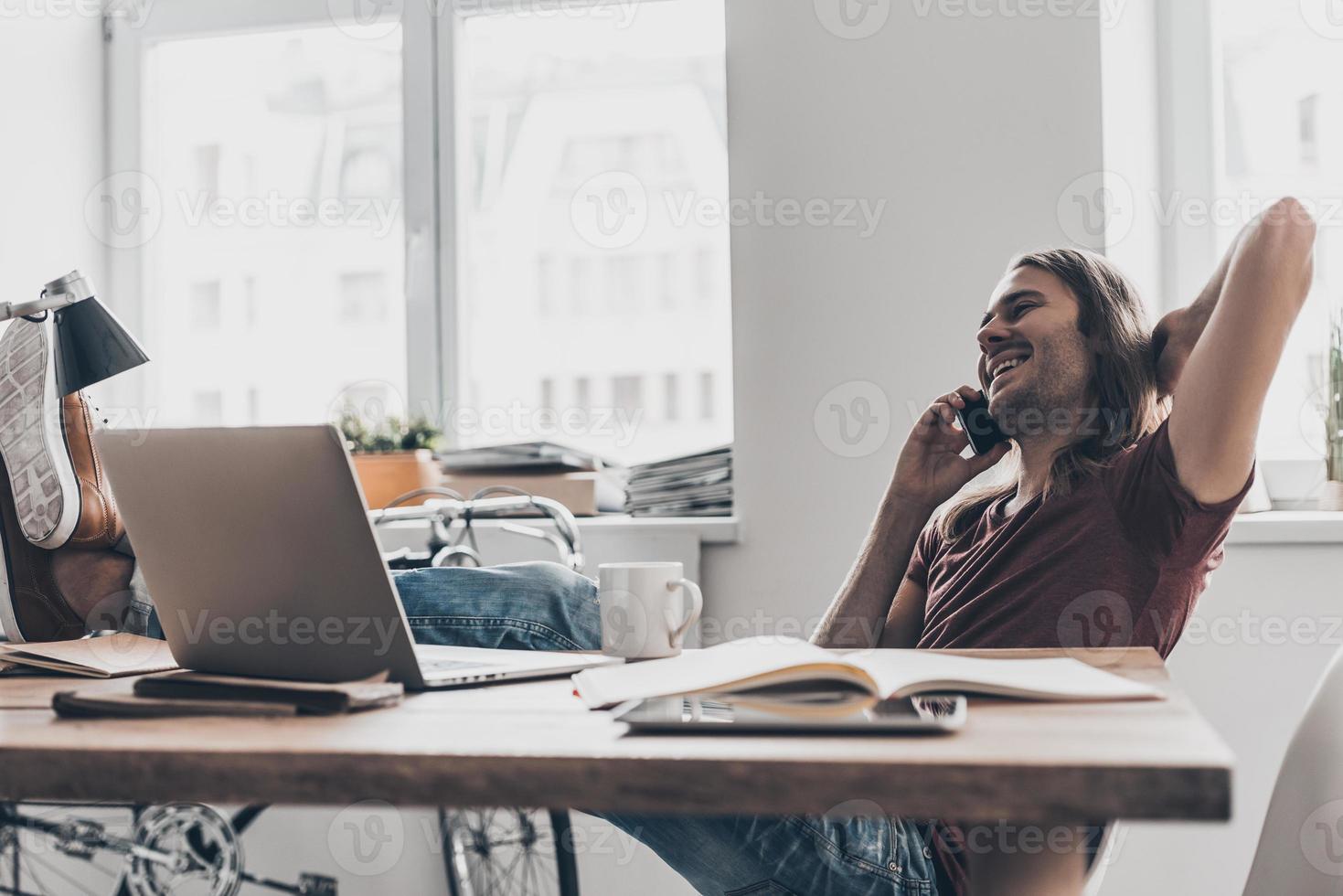 Amazing news Handsome young man with long hair talking on the mobile phone and looking happy while sitting at his working place in office photo