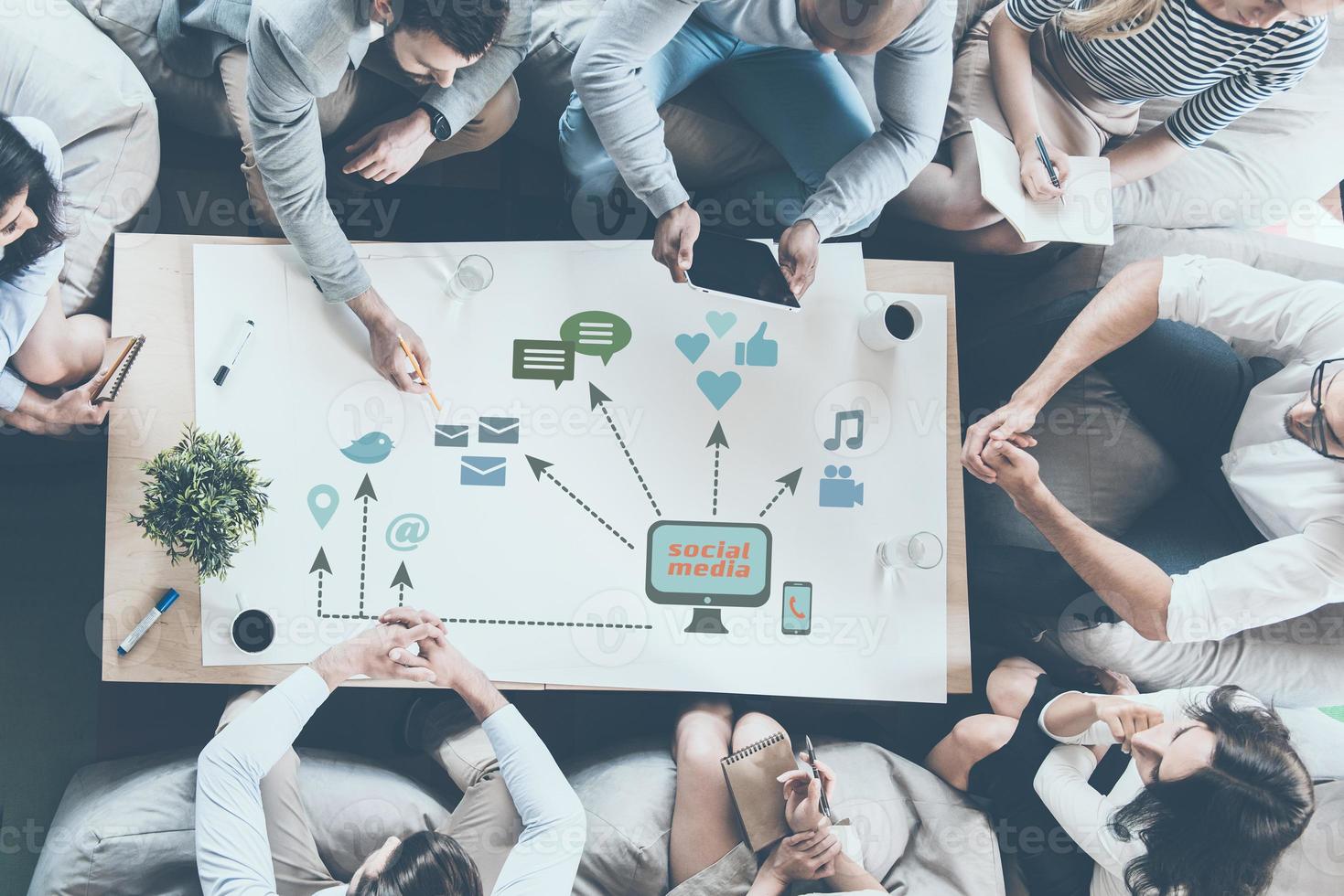 Discussing new strategy. Top view of business people discussing something while sitting around the desk together and pointing large paper with conceptual business icons drawn on it photo