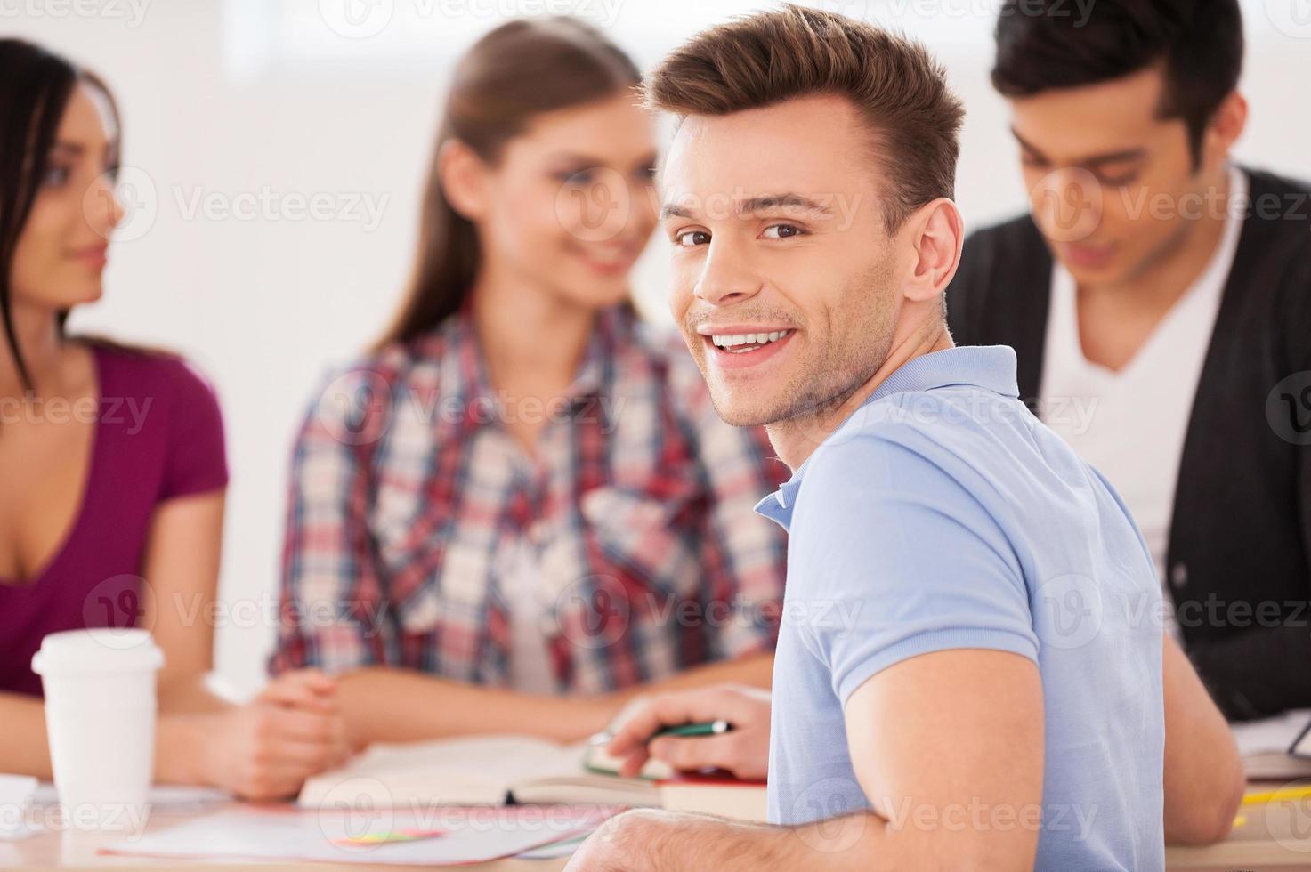 Feeling confident about his final exam. Four cheerful students sitting together at the desk and studying while one man looking over shoulder and smiling photo