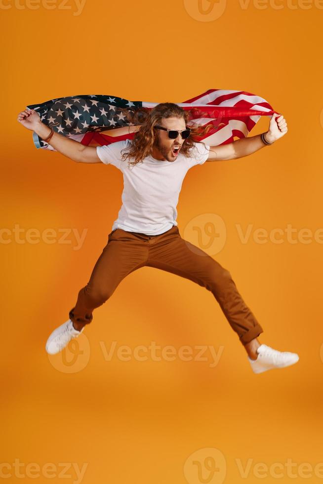 Confident young man in casual clothing making a face and looking away while holding American flag against yellow background photo