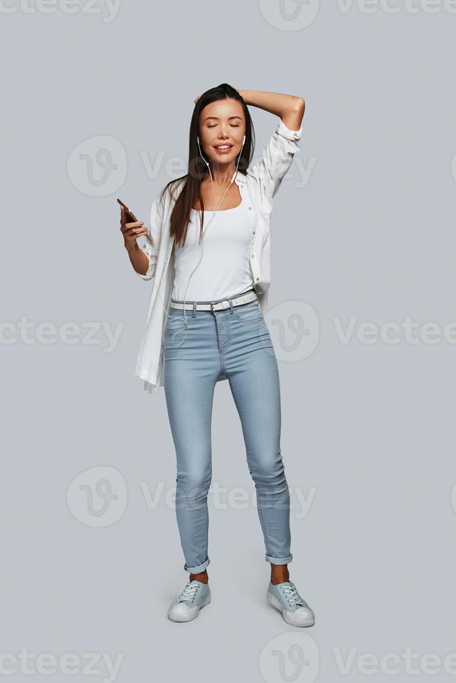 In love with music. Full length of young Asian woman dancing and smiling while standing against grey background photo