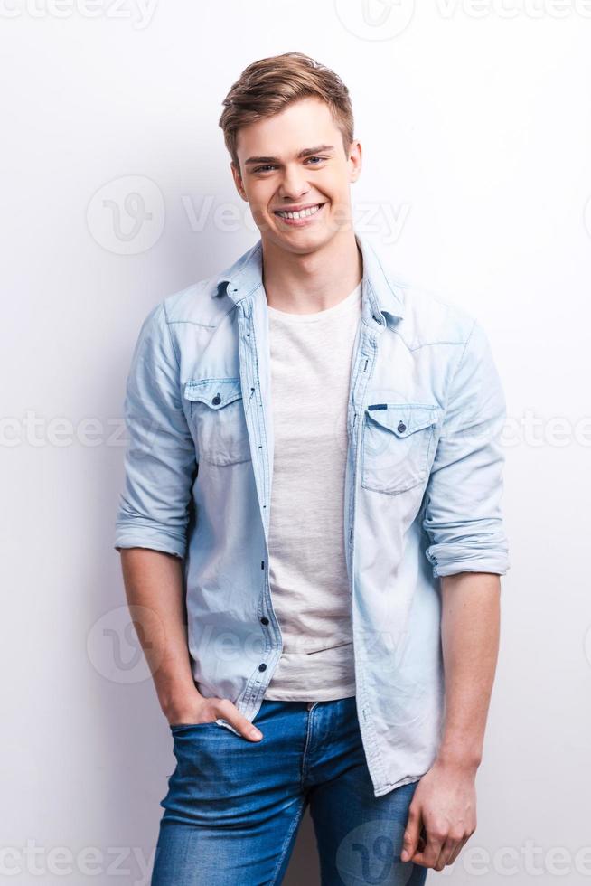 Casual lifestyle. Handsome young man holding hand in pocket and smiling while standing against grey background. photo