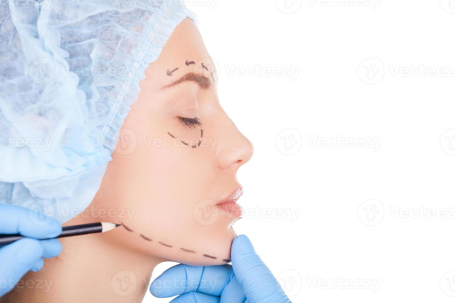 preparación del paciente para la cirugía. vista lateral de una hermosa joven con sombreros médicos manteniendo los ojos cerrados mientras el doctor esboza su rostro aislado en blanco foto