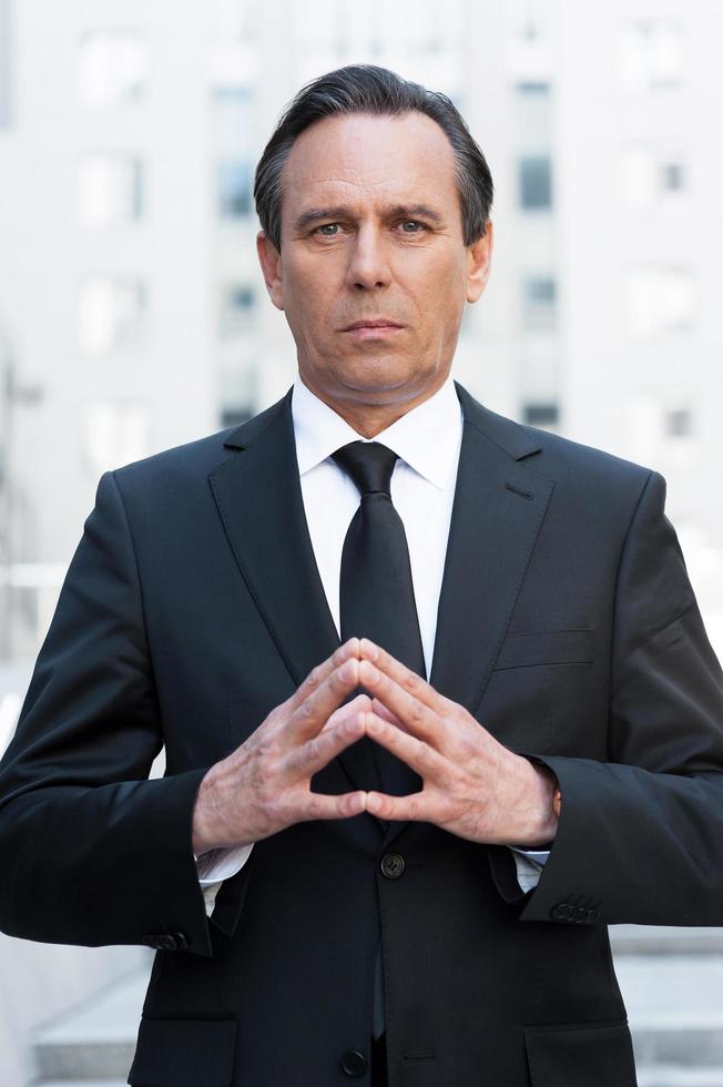 Power and success. Confident mature man in formalwear holding hands clasped and looking at camera while standing outdoors photo