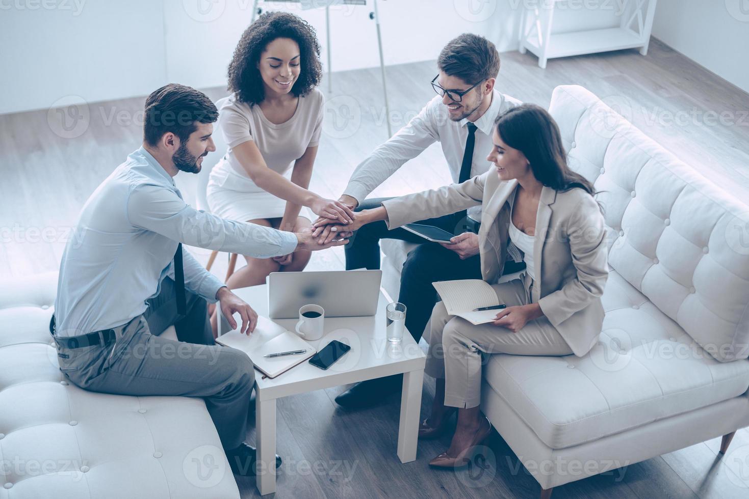 Ready for new achievements. Top view of group of four young people holding hands and showing their unity with smile while sitting on the couch at office photo