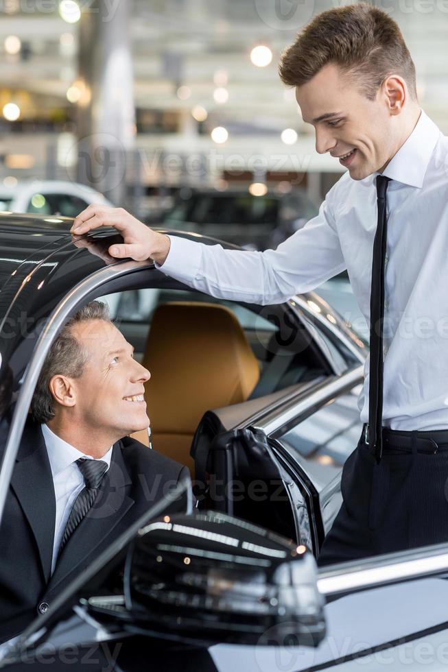 Let me show you all the features. Confident mature man ion formalwear sitting at the front seat of the car and talking to the car salesman standing near him photo