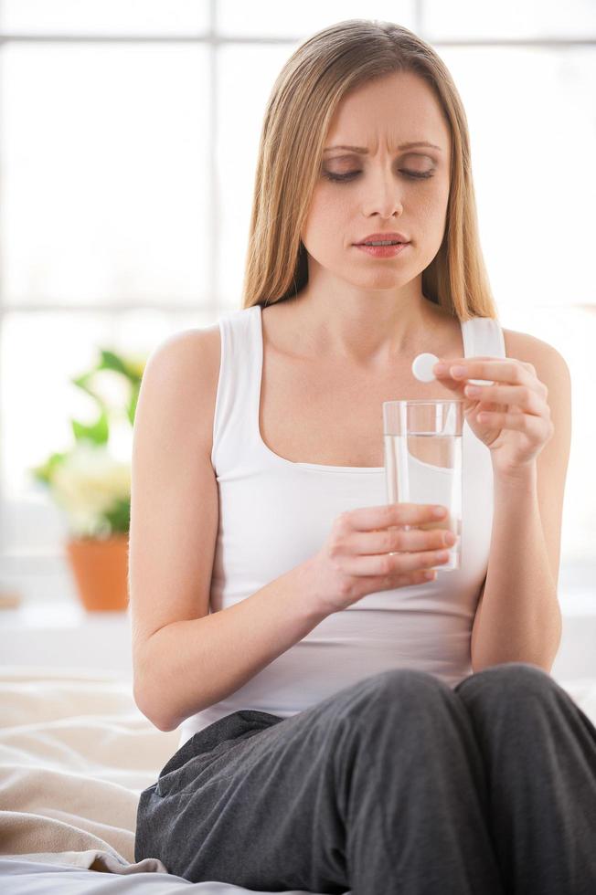 Woman taking aspirin. Young woman holding pill upon glass with water and expressing negativity while sitting in bed at her apartment photo