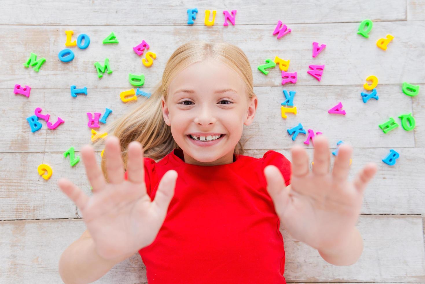 Having fun. Top view of cute little girl stretching out hands and smiling while lying on the floor with plastic colorful letters laying around her photo