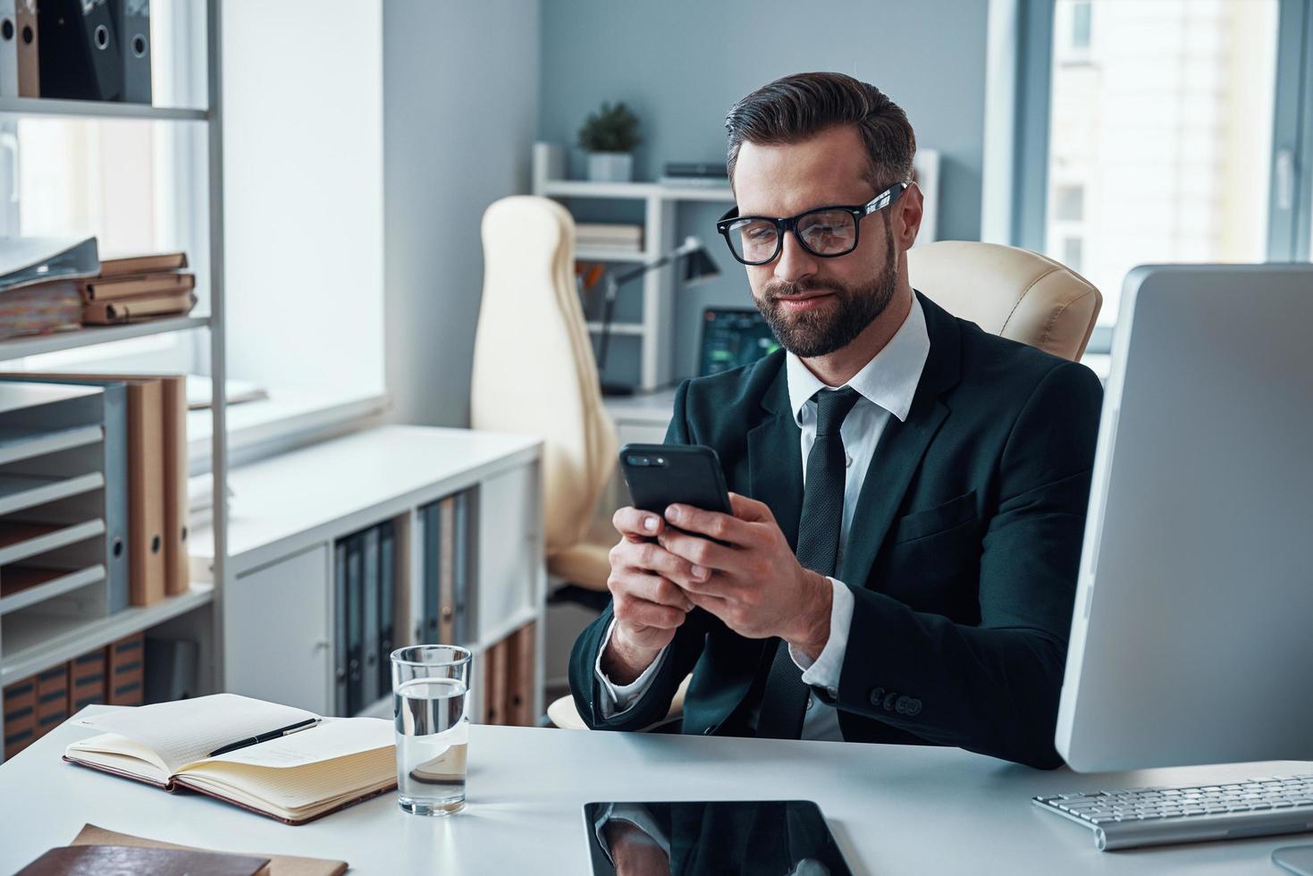 Good looking young man in shirt and tie using smart phone and smiling while sitting in the office photo