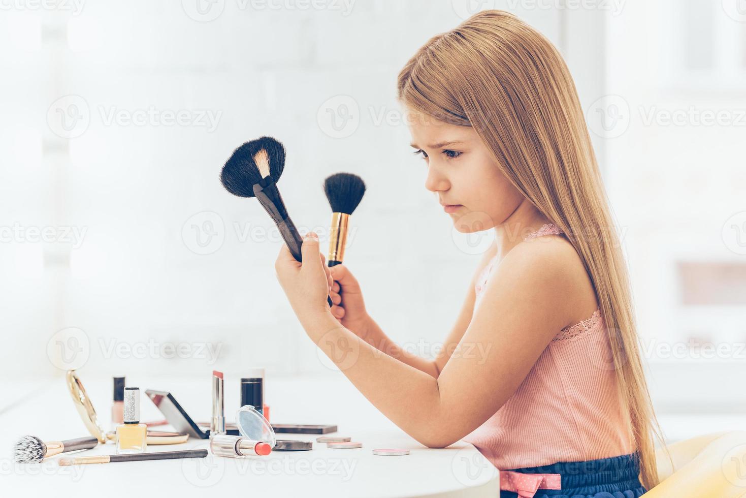 es este cepillo para contornear la vista lateral de una niña pensativa que elige uno de los dos cepillos de maquillaje mientras se sienta en el tocador foto