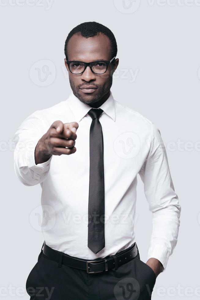 You are the one we need Handsome African man in eyeglasses pointing you while standing against grey background photo