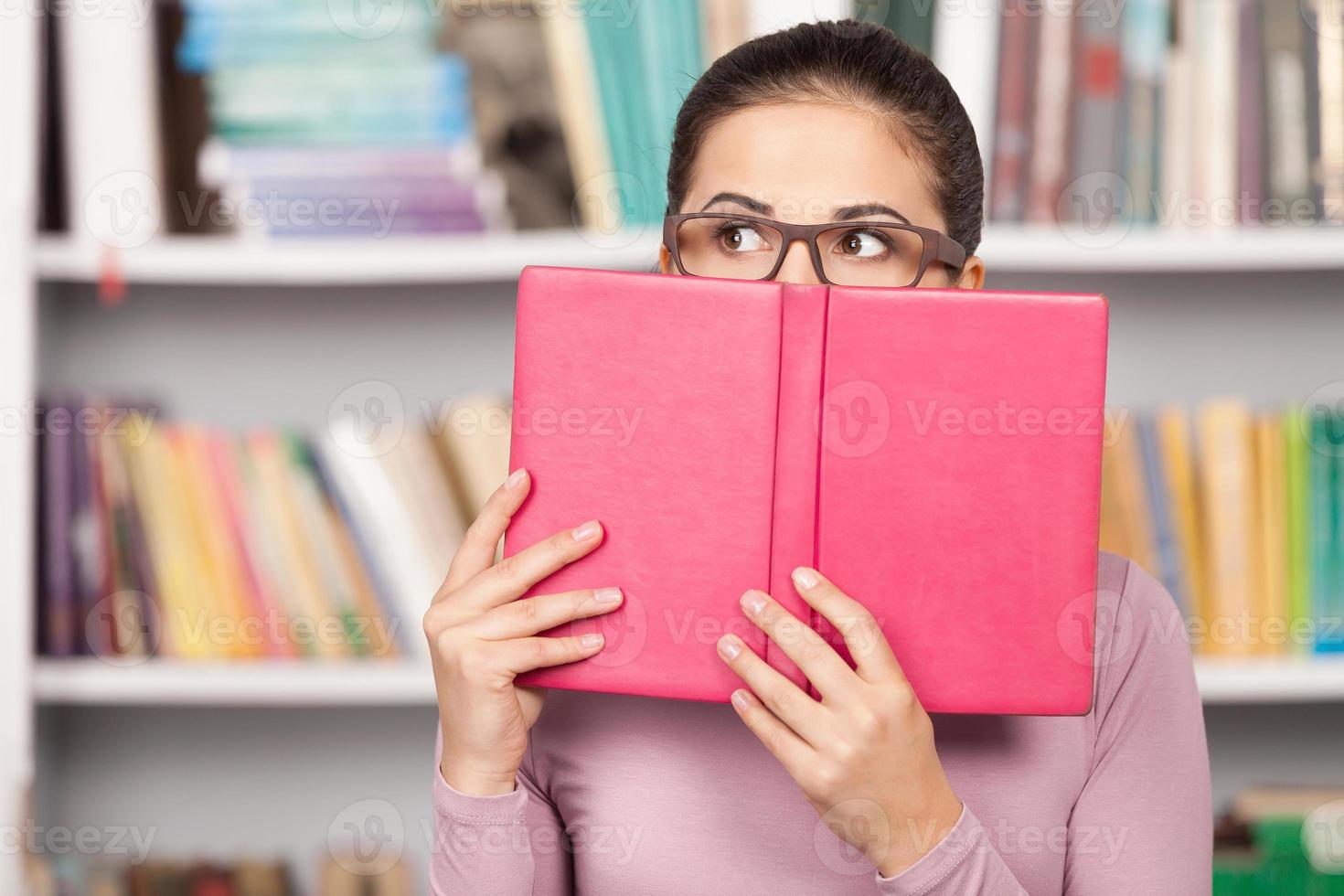 Worried about her exams. Terrified young woman looking out of the book while standing in front of the book shelf photo