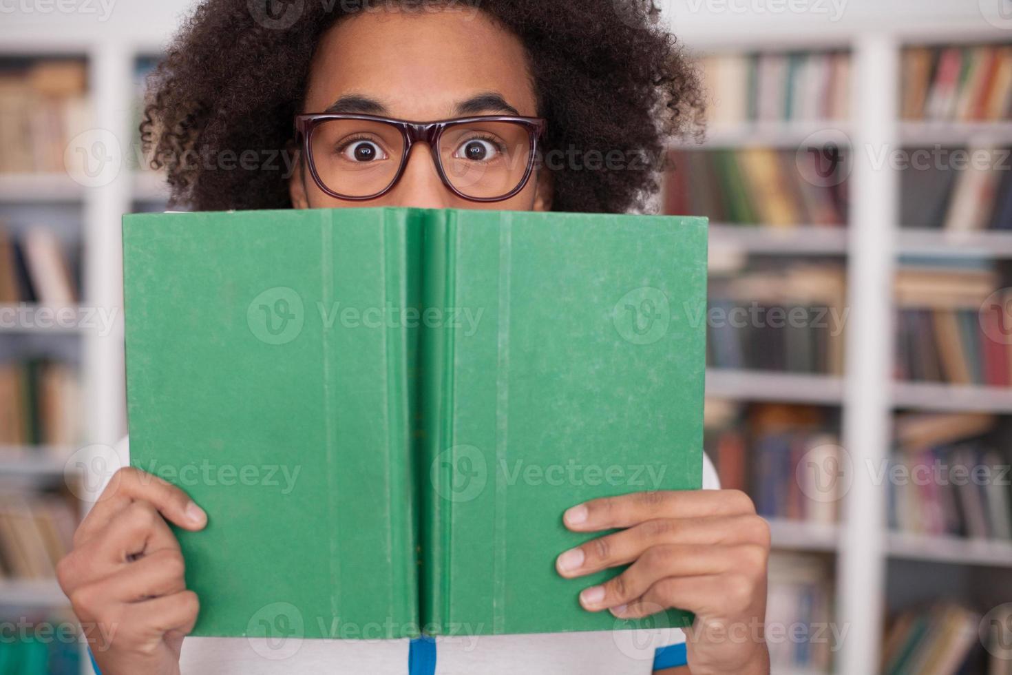 Worried about his exams. Terrified African teenager looking out of the book while standing in front of the book shelf photo