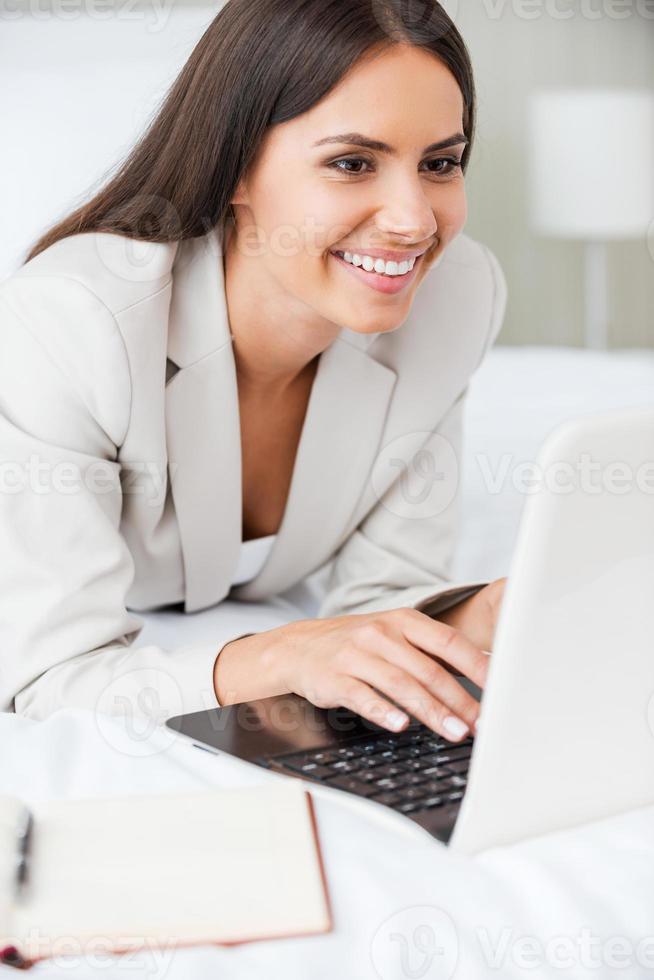 Staying in touch with colleagues in office. Confident young businesswoman in suit working on laptop and smiling while lying in bed at the hotel room photo