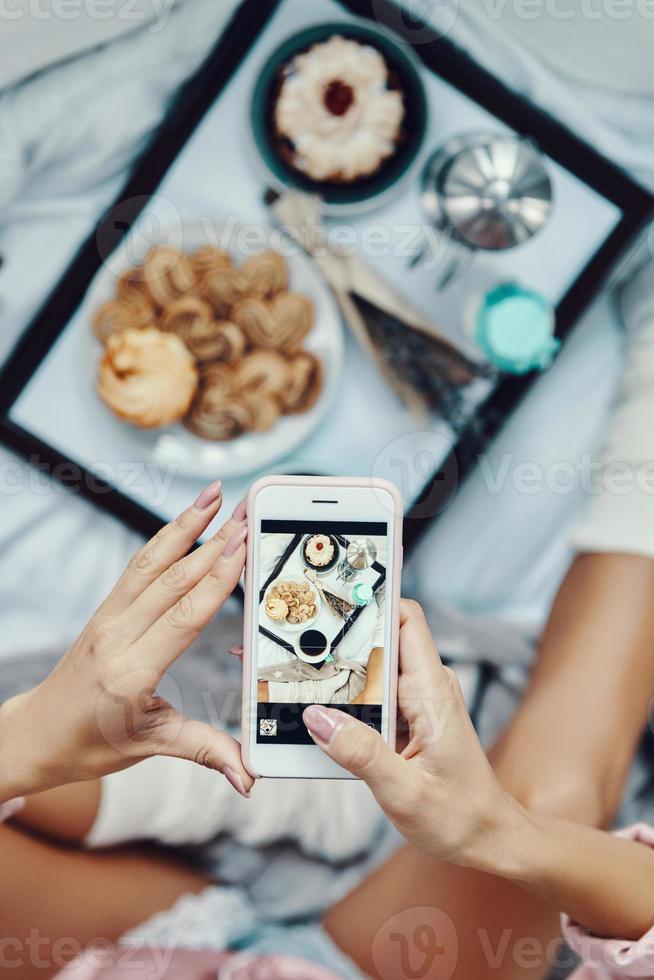 Close up top view of young woman in pajamas photographing flat lay of breakfast while resting in bed at home photo