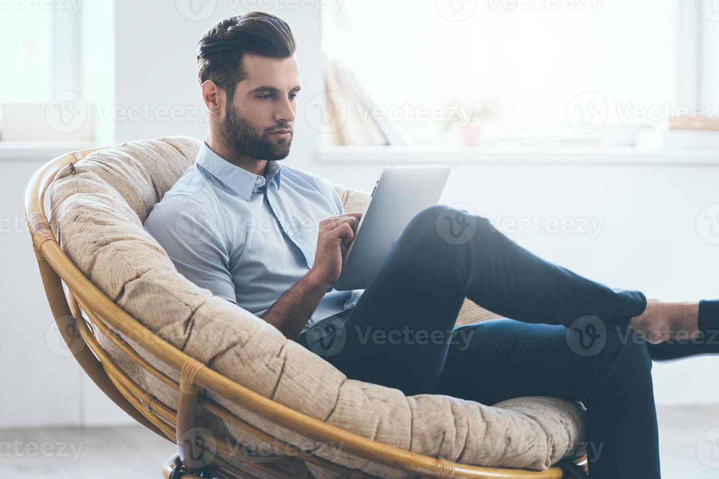 examinando su nueva tableta. un joven apuesto y confiado que trabaja en una tableta digital mientras se sienta en una silla grande y cómoda en casa foto