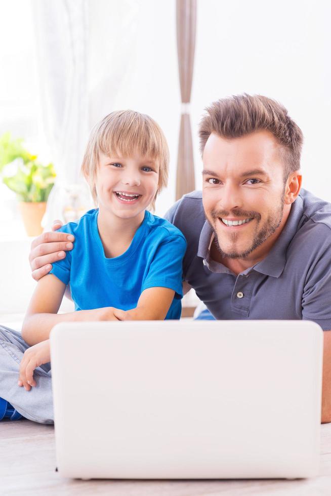Spending their free time together. Happy father embracing his cheerful little son while both sitting on the floor with laptop near them photo