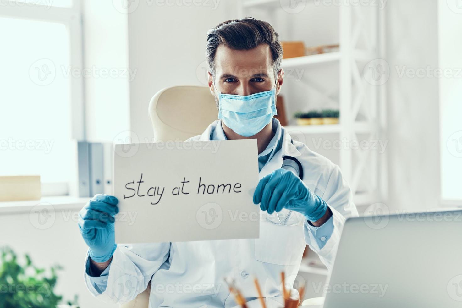 Young man in white lab coat and protective mask showing stay at home saying written on the paper while sitting indoors photo