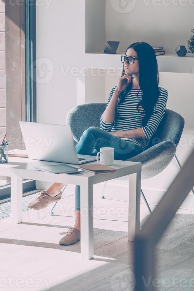 Lost in business thoughts. Thoughtful young beautiful woman holding hand on chin and looking through a window while sitting at her working place photo