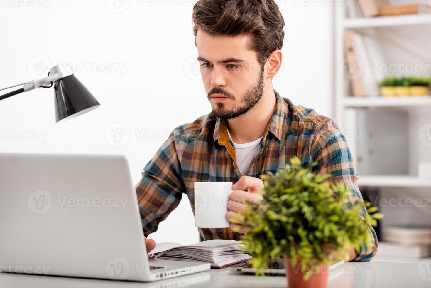 asegurándose de que toda la tarea se realice. un joven confiado que trabaja en una laptop y sostiene una taza de café mientras se sienta en su lugar de trabajo foto