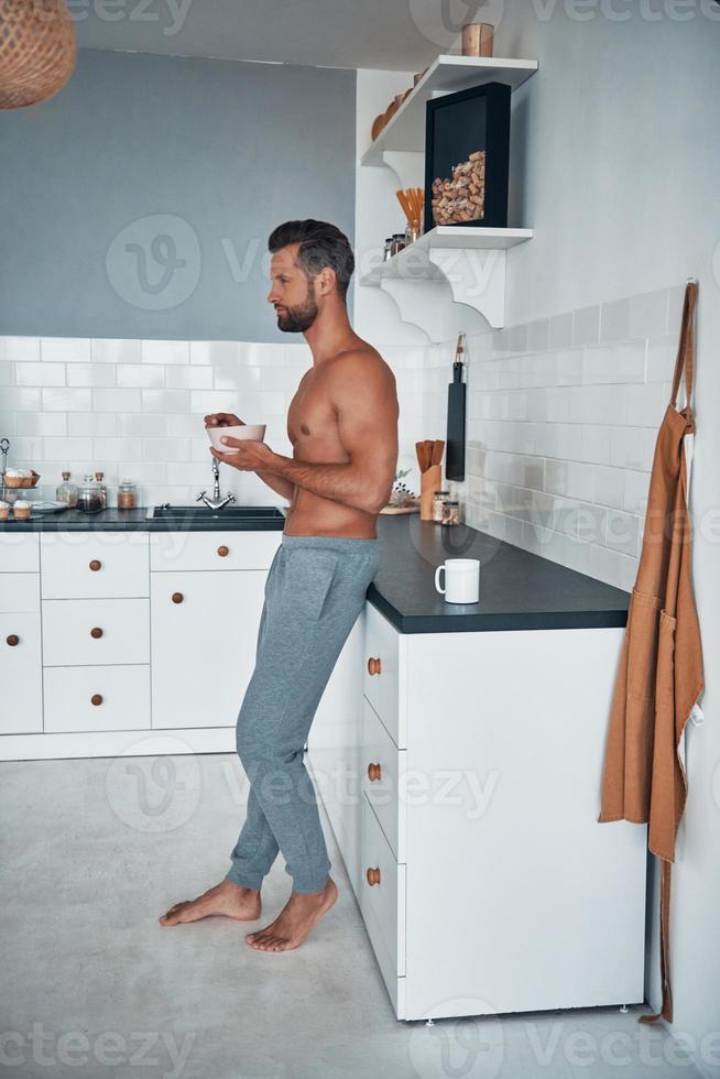 Full length of young shirtless man having breakfast while standing at home kitchen photo