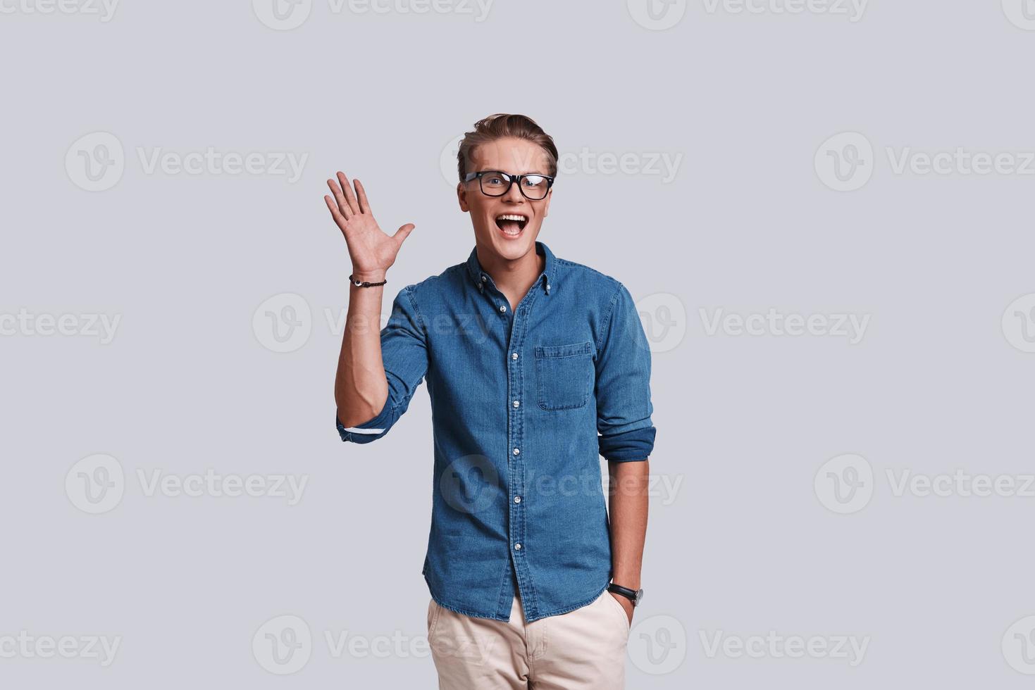 Good looking young man waving and smiling while standing against grey background photo