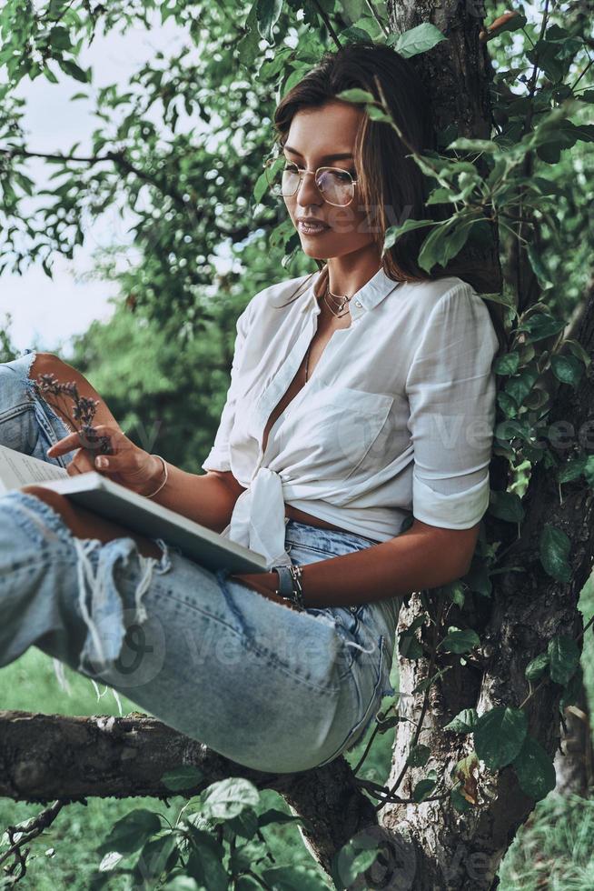 Perfect place to read. Attractive young woman in casual wear reading a book while sitting on the tree outdoors photo