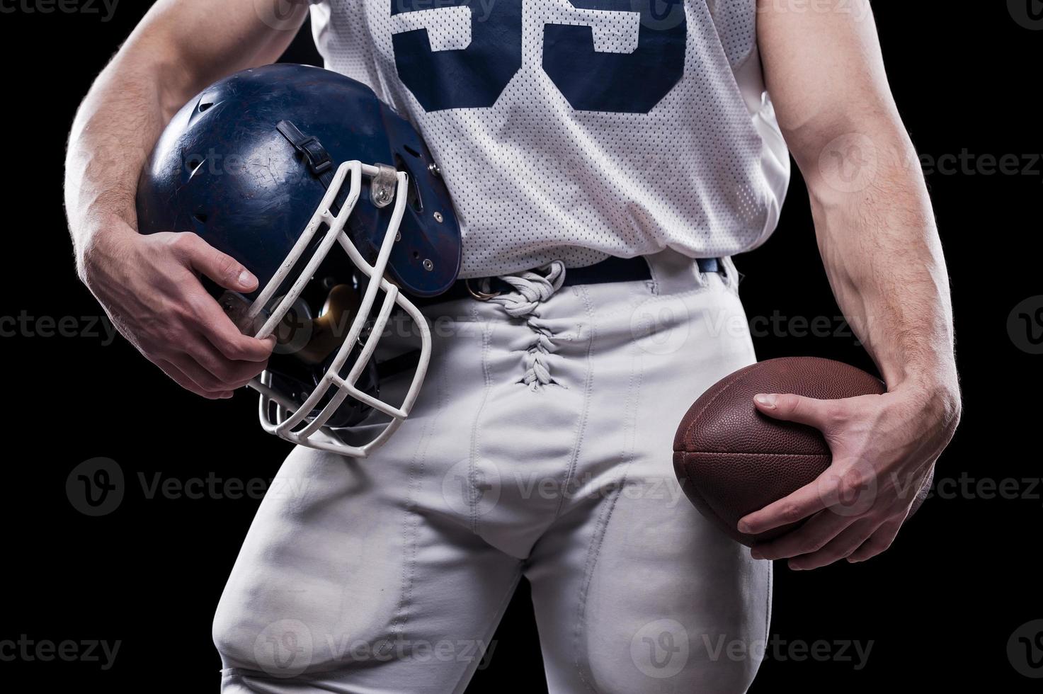 What is necessary for the game Cropped image of American football player holding ball and helmet while standing against black background photo