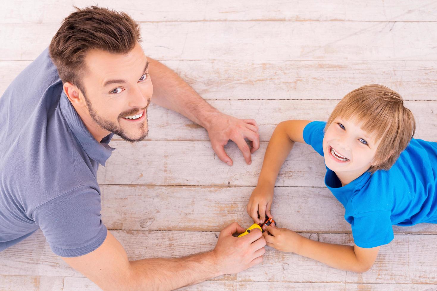 Playing together. Top view of happy father and son playing toy cars and looking up with smile while lying on the hardwood floor together photo