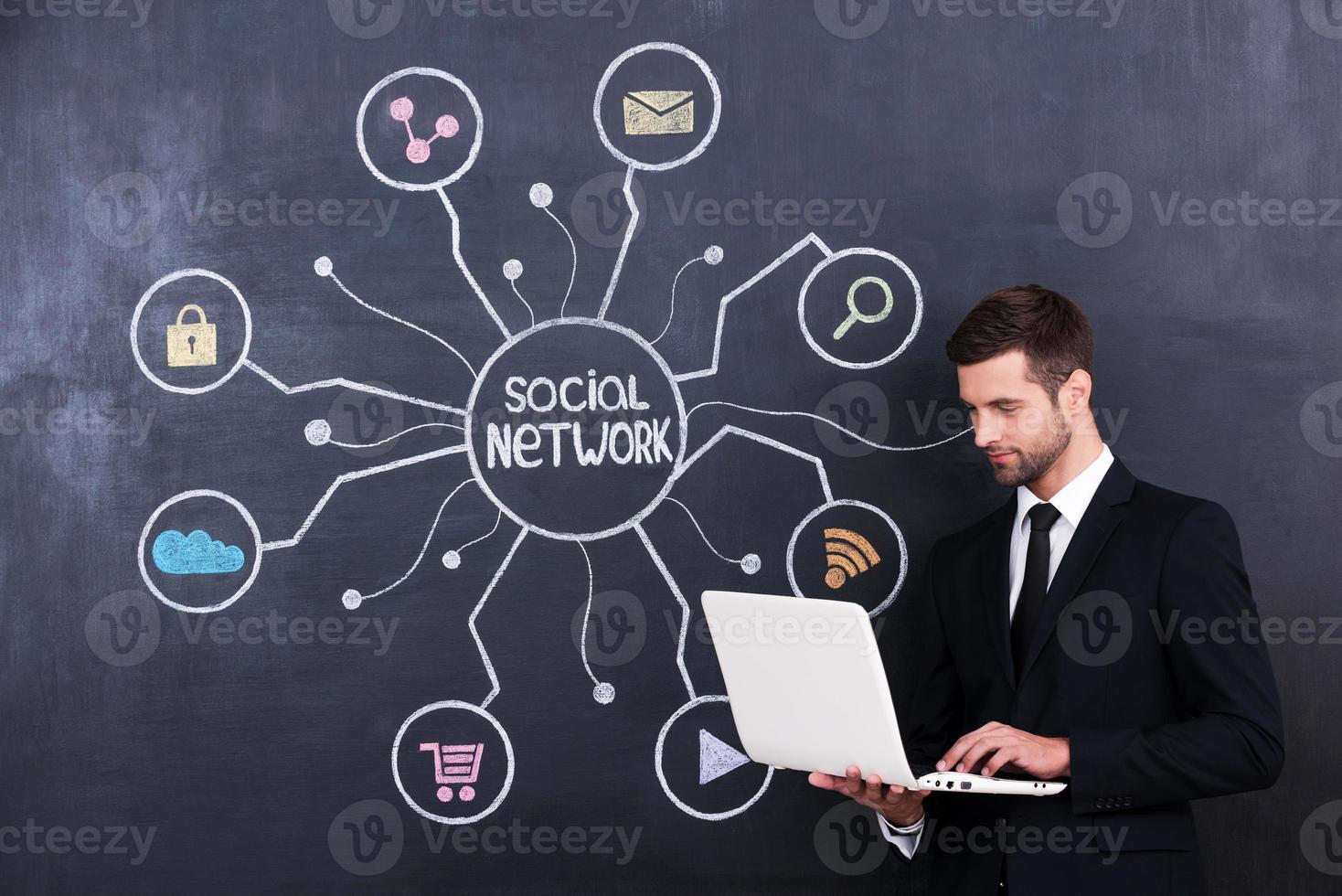 Social networking. Handsome young man working on laptop while standing against social network chalk drawing on blackboard photo