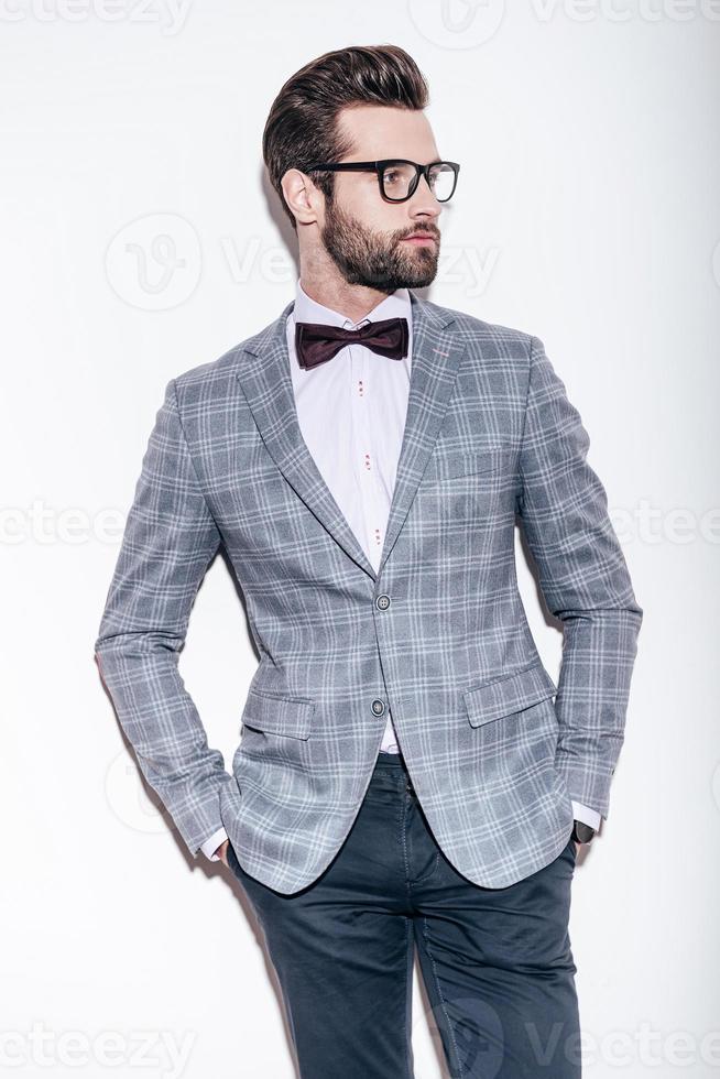 Style and intelligence. Handsome young man wearing suit and glasses keeping hands in pockets and looking away while standing against white background photo