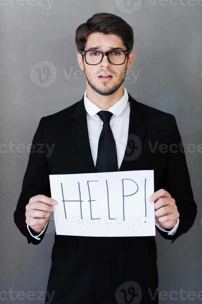 Asking for help. Frustrated young businessman holding paper with text and looking at camera while standing against grey background photo