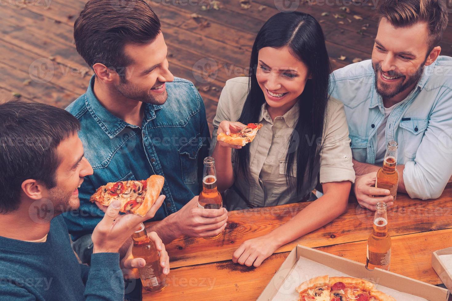 hora de pizza. vista superior de cinco personas alegres sosteniendo botellas con cerveza y comiendo pizza mientras están de pie al aire libre foto