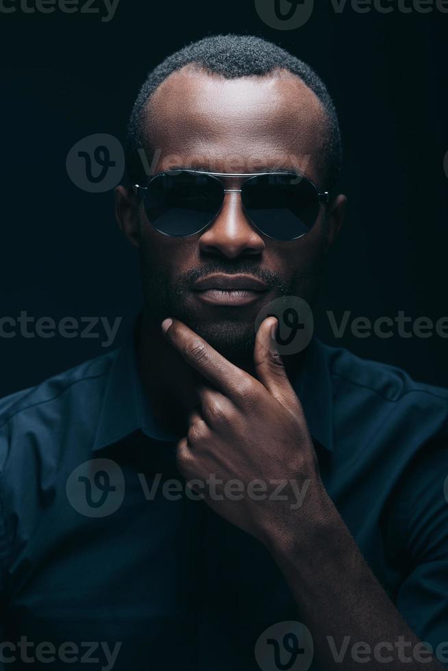Trendy look. Portrait of handsome young African man in sunglasses holding hand on chin and looking at camera while being in front of black background photo