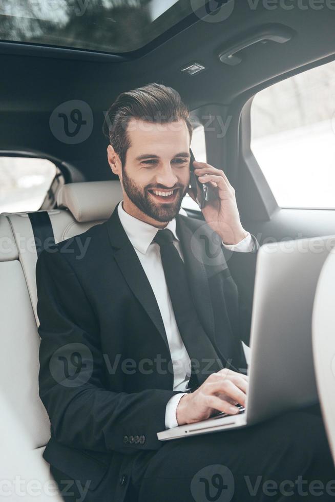 Great solutions every day. Handsome young businessman working on his laptop and talking on the phone while sitting in the car photo