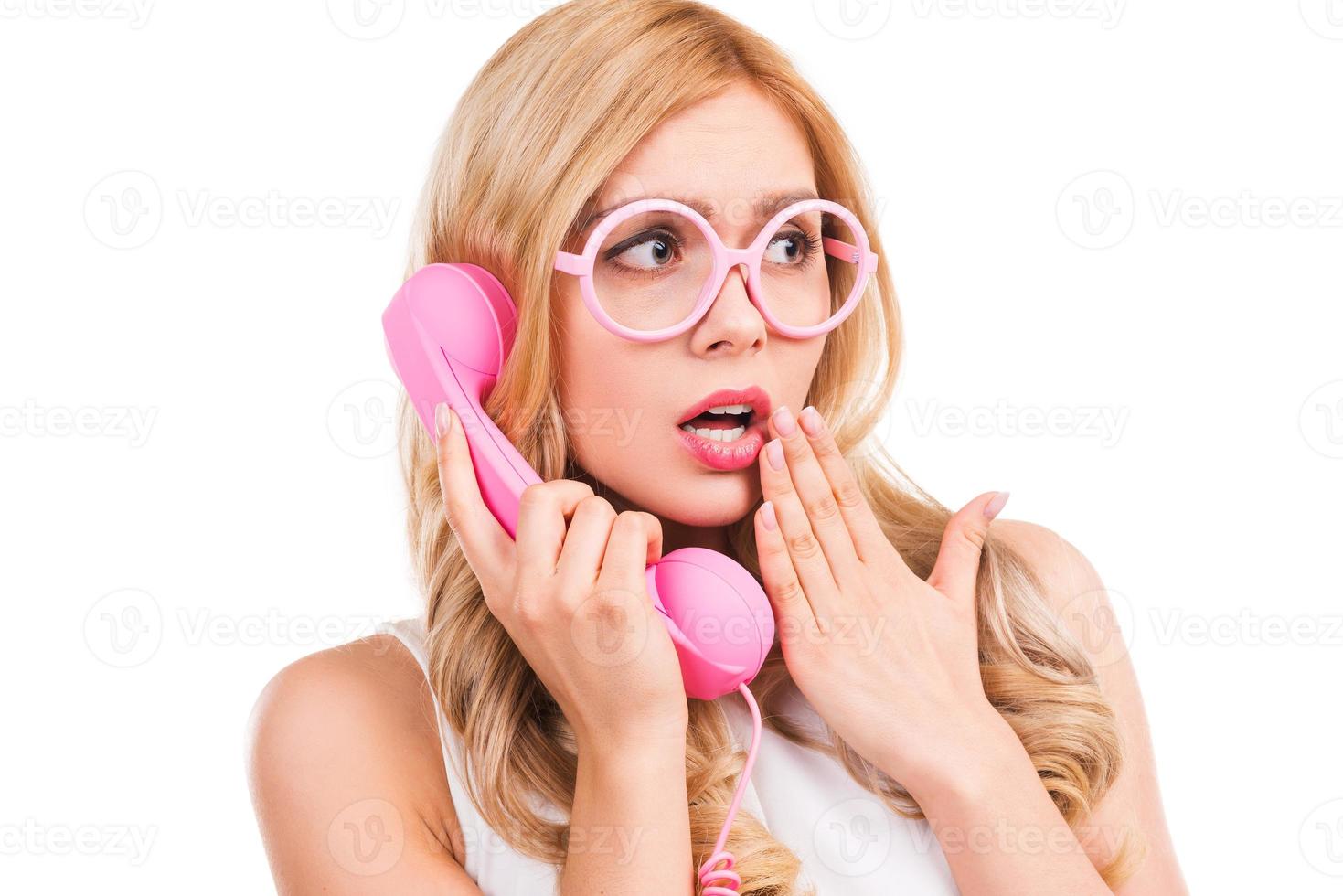 That is unbelievable Frustrated young blond hair woman talking on the telephone and covering mouth with hand while standing isolated on white background photo