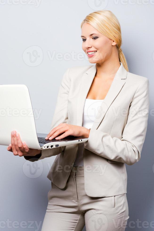 In love with her work. Confident young businesswoman holding laptop while standing against grey background photo