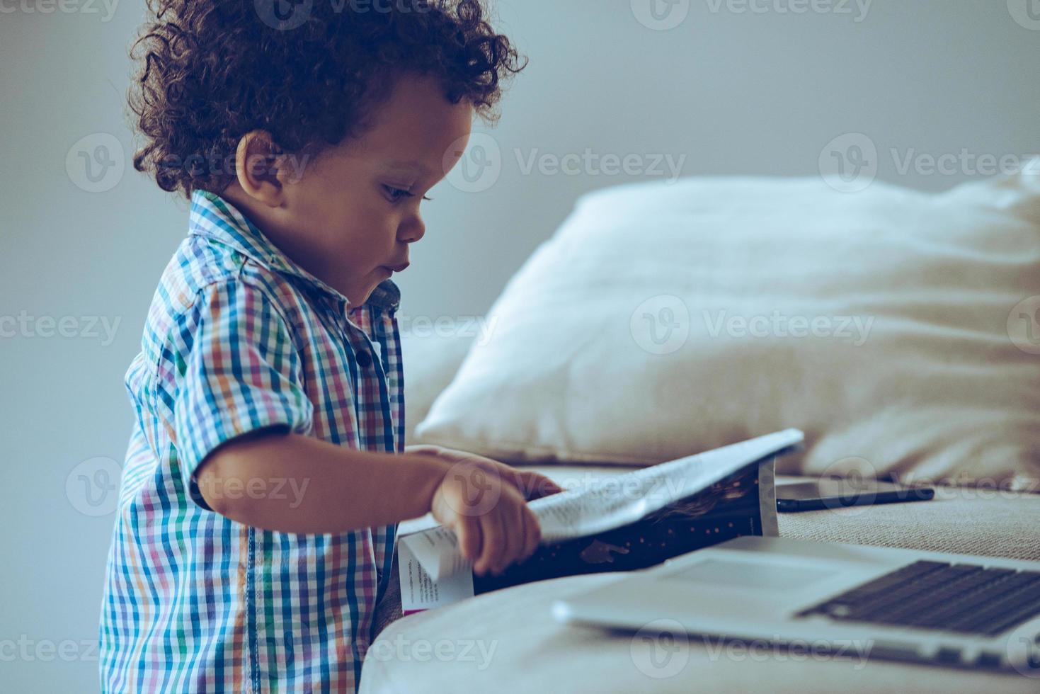 Looking through magazine. Side view of little African baby boy looking through magazine while standing near the couch at home photo