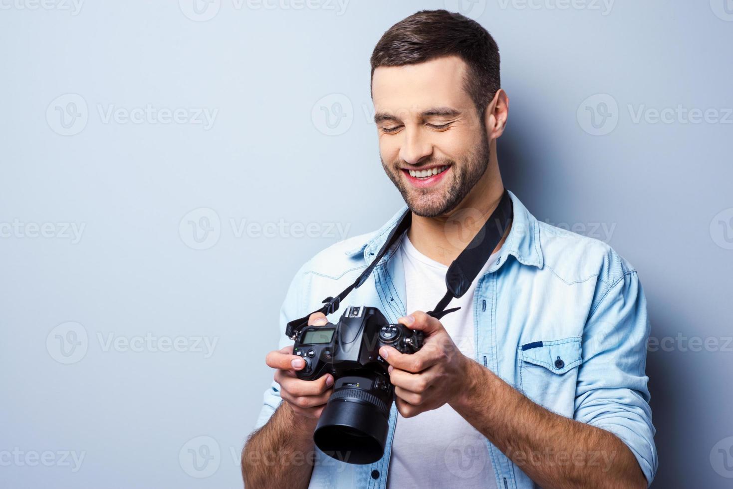 gran tiro guapo joven sosteniendo una cámara digital y mirándola con una sonrisa mientras está de pie contra el fondo gris foto