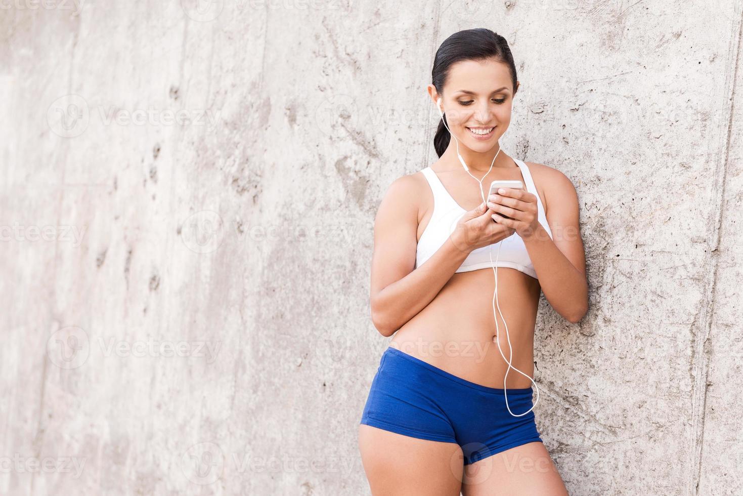 elegir la música adecuada para el entrenamiento. bella joven vestida con ropa deportiva sosteniendo un reproductor de mp3 y mirándolo con una sonrisa mientras se apoya en la pared de hormigón foto