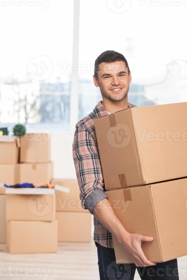 Moving to a new place. Cheerful young man holding a cardboard box and smiling at camera while more carton boxes laying on background photo