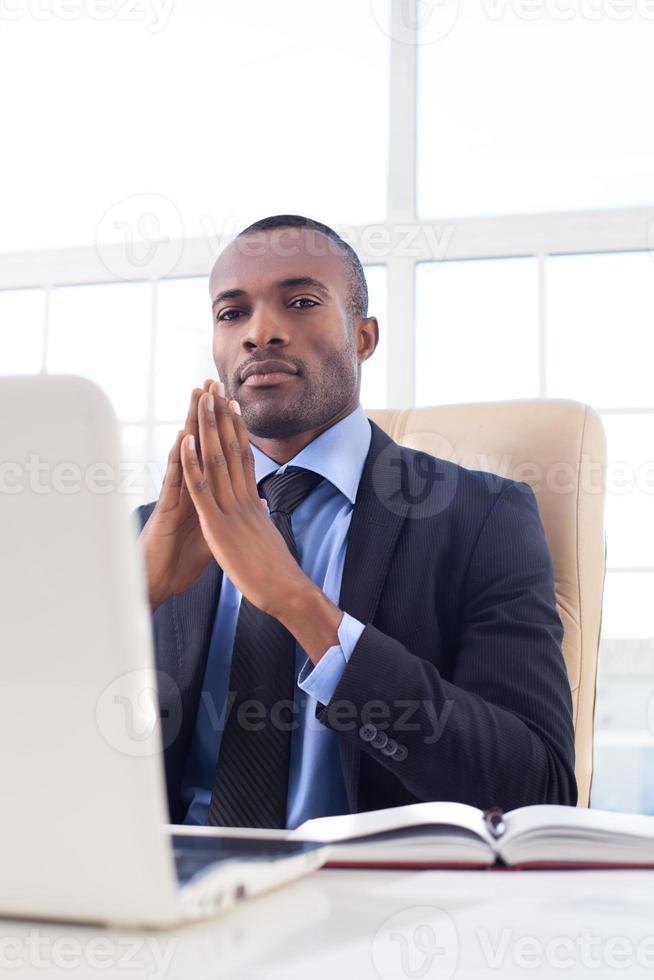 Confident in his power. Confident young African businessman keeping his hands clasped and looking at camera while sitting at his working place photo