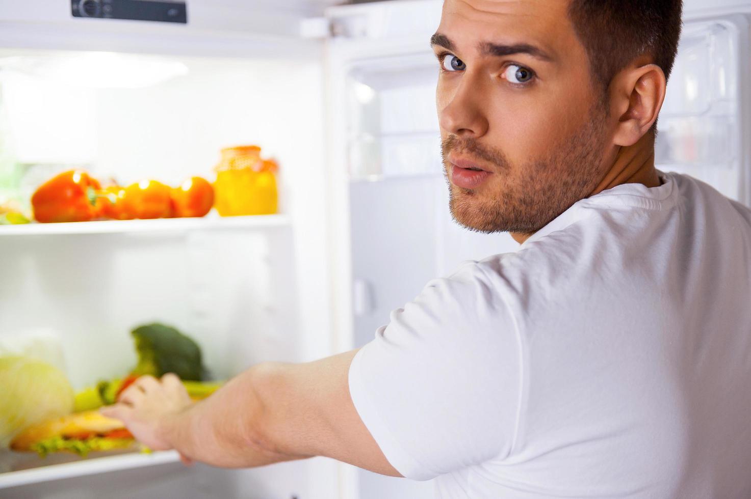 Feeling so hungry. Surprised young man standing near the open fridge and stretching hand to sandwich laying inside photo