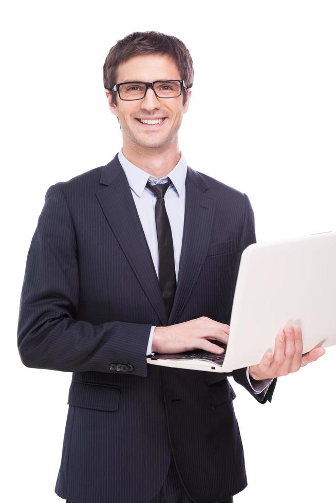 Quality support for your business. Handsome young man in formalwear working on laptop and smiling while standing isolated on white background photo