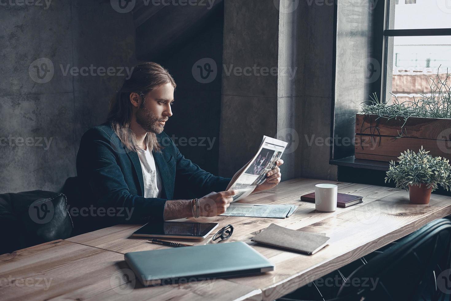Reading latest news. Handsome young man in smart casual wear reading a newspaper while sitting at his desk in office photo