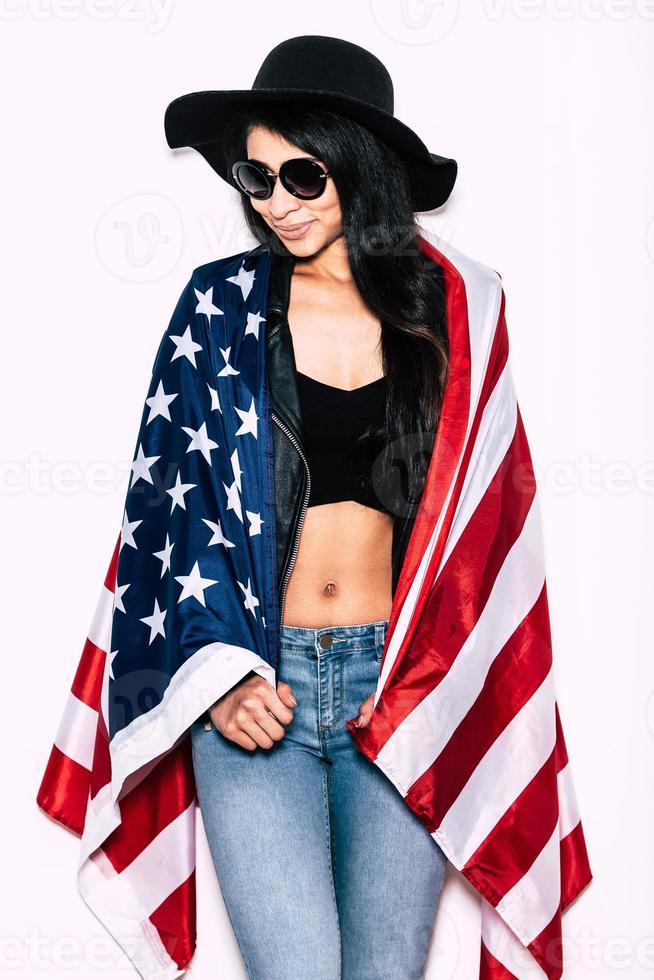Young and free. Beautiful young mixed race woman carrying American flag on shoulders and looking away with smile while standing against white background photo