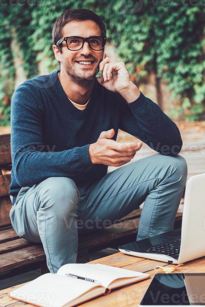 Good business talk. Happy young man talking on the mobile phonewhile sitting at the wooden table outdoors photo