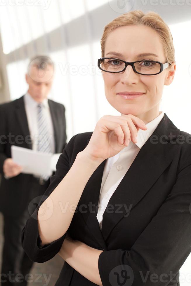 confiado e inteligente. atractiva mujer de negocios madura sosteniendo la mano en la barbilla y sonriendo a la cámara mientras el hombre en ropa formal está de pie en el fondo foto