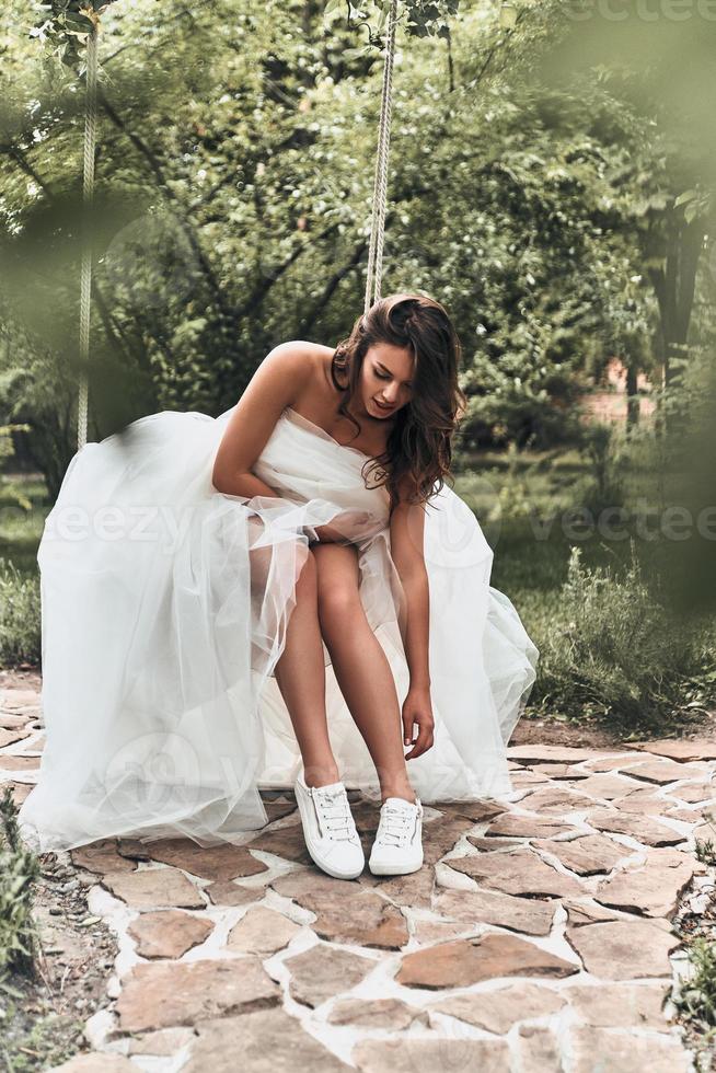 Beautiful modern bride. Attractive young woman in wedding dress adjusting her sports shoes while sitting on the swing outdoors photo